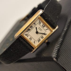 May include: A rectangular Seiko Quartz watch with a gold-toned case and a black leather strap. The watch face is white with black Roman numerals and hands. The watch is displayed on a textured black surface.