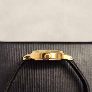 K&ouml;nnte beinhalten: Eine goldfarbene Armbanduhr mit einem schwarzen Lederarmband. Das Zifferblatt der Uhr ist rund und hat eine goldene L&uuml;nette. Das Uhrenarmband ist d&uuml;nn und scheint aus Leder zu sein. Die Uhr wird von der Seite betrachtet.