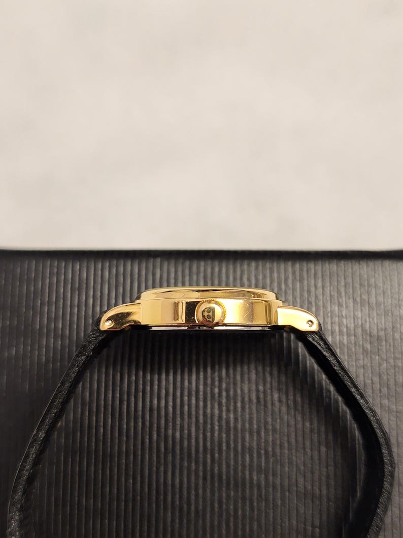 K&ouml;nnte beinhalten: Eine goldfarbene Armbanduhr mit einem schwarzen Lederarmband. Das Zifferblatt der Uhr ist rund und hat eine goldene L&uuml;nette. Das Uhrenarmband ist d&uuml;nn und scheint aus Leder zu sein. Die Uhr wird von der Seite betrachtet.