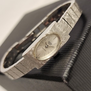 May include: A silver Gruen watch with a white face and a silver band. The watch face has the words "Gruen Precision" printed on it.