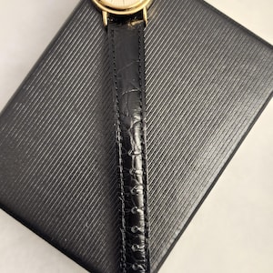 K&ouml;nnte beinhalten: Eine goldfarbene Lorus-Armbanduhr mit schwarzem Lederarmband liegt auf einer strukturierten schwarzen Oberfl&auml;che. Das Zifferblatt der Uhr ist hellgoldfarben mit schwarzen Zeigern und Markierungen. Das Uhrenarmband ist strukturiert und scheint aus Leder zu sein.