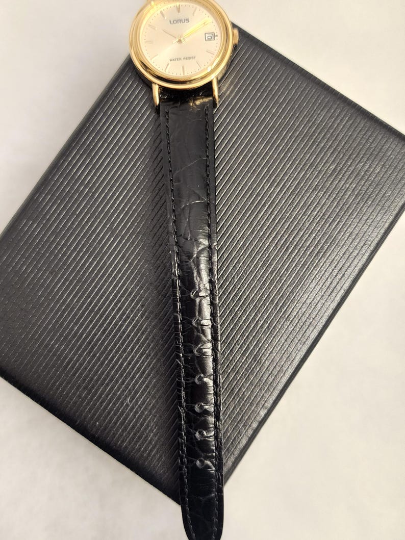 K&ouml;nnte beinhalten: Eine goldfarbene Lorus-Armbanduhr mit schwarzem Lederarmband liegt auf einer strukturierten schwarzen Oberfl&auml;che. Das Zifferblatt der Uhr ist hellgoldfarben mit schwarzen Zeigern und Markierungen. Das Uhrenarmband ist strukturiert und scheint aus Leder zu sein.