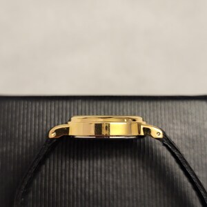 K&ouml;nnte beinhalten: Eine goldfarbene Armbanduhr mit einem schwarzen Lederarmband. Das Uhrengeh&auml;use ist rund und hat eine polierte Oberfl&auml;che. Das schwarze Armband ist d&uuml;nn und scheint aus Leder zu sein. Die Uhr ist vor einem dunklen, strukturierten Hintergrund platziert.