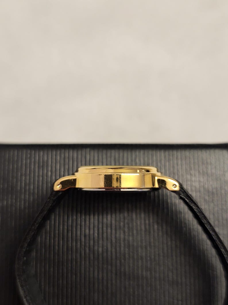 K&ouml;nnte beinhalten: Eine goldfarbene Armbanduhr mit einem schwarzen Lederarmband. Das Uhrengeh&auml;use ist rund und hat eine polierte Oberfl&auml;che. Das schwarze Armband ist d&uuml;nn und scheint aus Leder zu sein. Die Uhr ist vor einem dunklen, strukturierten Hintergrund platziert.