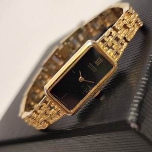 Peut inclure: Une montre Seiko Quartz dorée avec un cadran rectangulaire noir, des aiguilles dorées et un chiffre romain à douze heures. La montre est dotée d'un bracelet en métal doré texturé de style maillons avec un motif répétitif.