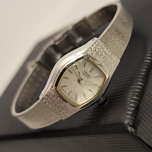 May include: A silver Seiko Quartz watch with a textured silver strap. The watch face is silver with black hands and markers. The watch has a rectangular face with rounded edges. The watch is displayed on a black surface.