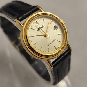 K&ouml;nnte beinhalten: Goldfarbene Lorus-Uhr mit cremefarbenem Zifferblatt und schwarzem Lederarmband. Die Uhr hat eine Datumsanzeige und den Schriftzug "WATER RESIST" auf dem Zifferblatt. Die Uhr hat goldene Zeiger und Stundenmarkierungen.