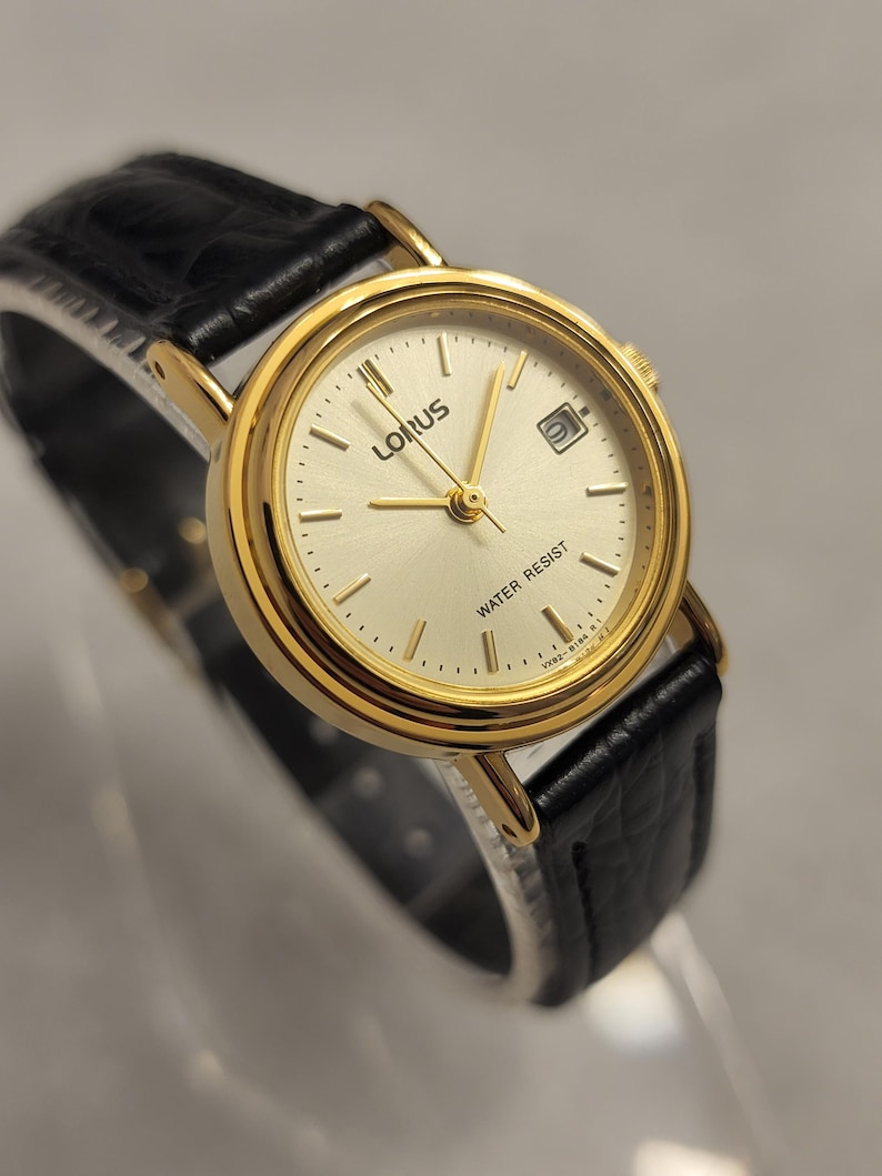 K&ouml;nnte beinhalten: Goldfarbene Lorus-Uhr mit cremefarbenem Zifferblatt und schwarzem Lederarmband. Die Uhr hat eine Datumsanzeige und den Schriftzug "WATER RESIST" auf dem Zifferblatt. Die Uhr hat goldene Zeiger und Stundenmarkierungen.