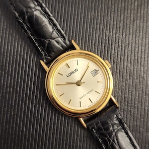 K&ouml;nnte beinhalten: Eine goldfarbene Lorus-Armbanduhr mit einem schwarzen Lederarmband. Das Zifferblatt der Uhr ist hell mit schwarzen Zeigern und Markierungen sowie einem Datumsfenster bei 3 Uhr. Das Armband ist etwa 2 cm breit.