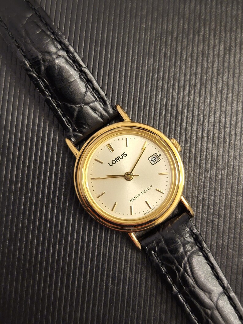 K&ouml;nnte beinhalten: Eine goldfarbene Lorus-Armbanduhr mit einem schwarzen Lederarmband. Das Zifferblatt der Uhr ist hell mit schwarzen Zeigern und Markierungen sowie einem Datumsfenster bei 3 Uhr. Das Armband ist etwa 2 cm breit.