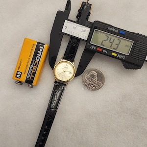 K&ouml;nnte beinhalten: Eine goldfarbene Lorus-Uhr mit schwarzem Lederarmband wird mit einem digitalen Messschieber gemessen, der 24,3 mm anzeigt. Eine gelbe Duracell-Batterie und eine M&uuml;nze sind ebenfalls im Bild. Die Uhr hat eine Datumsanzeige.