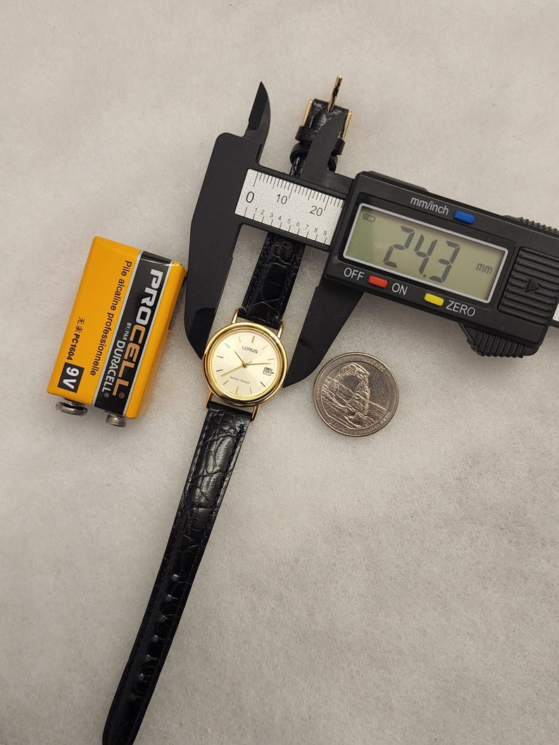 K&ouml;nnte beinhalten: Eine goldfarbene Lorus-Uhr mit schwarzem Lederarmband wird mit einem digitalen Messschieber gemessen, der 24,3 mm anzeigt. Eine gelbe Duracell-Batterie und eine M&uuml;nze sind ebenfalls im Bild. Die Uhr hat eine Datumsanzeige.