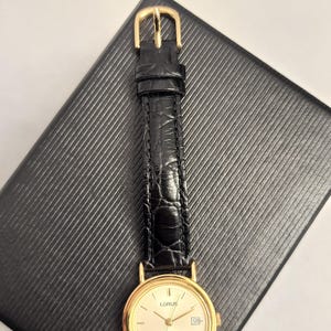 K&ouml;nnte beinhalten: Eine goldfarbene Lorus-Uhr mit cremefarbenem Zifferblatt und schwarzem Lederarmband. Die Uhr hat eine Datumsanzeige und liegt auf einer strukturierten schwarzen Oberfl&auml;che. Das Armband wird mit einer goldenen Schnalle befestigt.