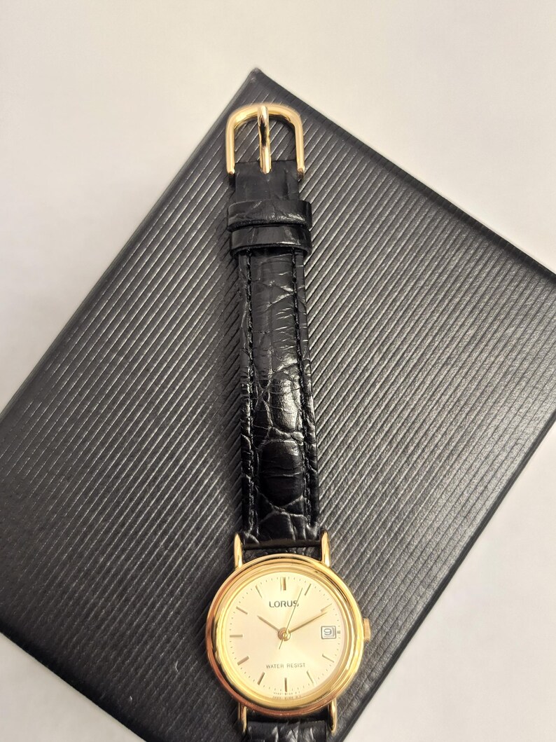 K&ouml;nnte beinhalten: Eine goldfarbene Lorus-Uhr mit cremefarbenem Zifferblatt und schwarzem Lederarmband. Die Uhr hat eine Datumsanzeige und liegt auf einer strukturierten schwarzen Oberfl&auml;che. Das Armband wird mit einer goldenen Schnalle befestigt.