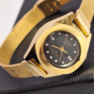 Peut inclure: Une montre-bracelet dorée avec un cadran noir et un bracelet en maille dorée. Le cadran de la montre a un mouvement à quartz et est chronométré électroniquement. Le cadran de la montre comporte également le texte "Longine Echo" et "Quartzomatic Electronically Timed" imprimé dessus.