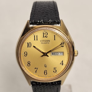 May include: A gold-toned Citizen Quartz wristwatch with a black leather strap. The watch face is gold with black numbers and hands. The date and day of the week are displayed in a small window at the 3 o'clock position. The watch face also has the text "CITIZEN QUARTZ" printed on it.