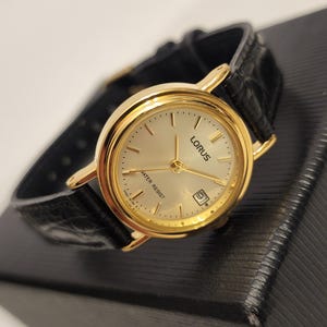 K&ouml;nnte beinhalten: Eine goldfarbene Lorus-Uhr mit cremefarbenem Zifferblatt und schwarzem Lederarmband. Die Uhr hat goldene Zeiger und Markierungen, eine Datumsanzeige und den Text "WATER RESIST". Die Uhr liegt auf einer schwarzen Oberfl&auml;che.