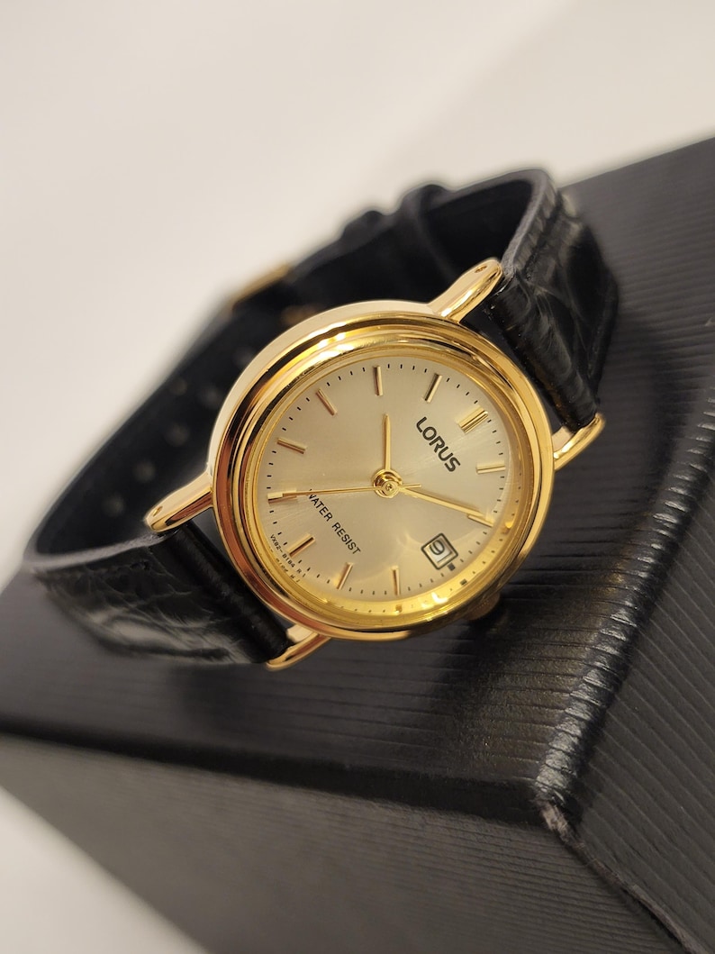 K&ouml;nnte beinhalten: Eine goldfarbene Lorus-Uhr mit cremefarbenem Zifferblatt und schwarzem Lederarmband. Die Uhr hat goldene Zeiger und Markierungen, eine Datumsanzeige und den Text "WATER RESIST". Die Uhr liegt auf einer schwarzen Oberfl&auml;che.