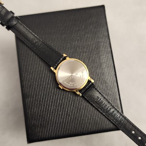 K&ouml;nnte beinhalten: Eine goldfarbene Lorus-Uhr mit schwarzem Lederarmband liegt auf einer schwarzen, strukturierten Schachtel. Das Zifferblatt der Uhr ist silberfarben, und das Armband ist mit dem Markennamen versehen. Die Uhr ist ein klassisches Design, geeignet f&uuml;r den Alltag.