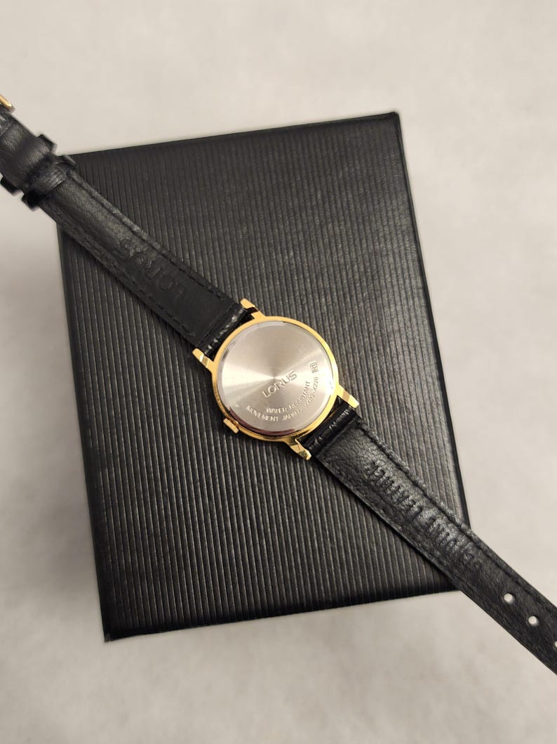 K&ouml;nnte beinhalten: Eine goldfarbene Lorus-Uhr mit schwarzem Lederarmband liegt auf einer schwarzen, strukturierten Schachtel. Das Zifferblatt der Uhr ist silberfarben, und das Armband ist mit dem Markennamen versehen. Die Uhr ist ein klassisches Design, geeignet f&uuml;r den Alltag.