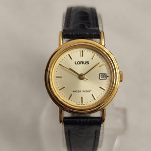 K&ouml;nnte beinhalten: Eine goldfarbene Lorus Armbanduhr mit schwarzem Lederarmband. Das Zifferblatt der Uhr ist hell champagnerfarben mit schwarzen Stundenmarkierungen und Zeigern. Die Uhr hat eine Datumsanzeige bei 3 Uhr und die Worte "WATER RESIST" sind auf dem Zifferblatt aufgedruckt.
