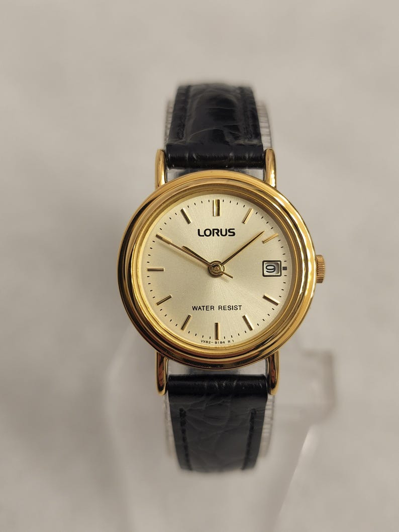 K&ouml;nnte beinhalten: Eine goldfarbene Lorus Armbanduhr mit schwarzem Lederarmband. Das Zifferblatt der Uhr ist hell champagnerfarben mit schwarzen Stundenmarkierungen und Zeigern. Die Uhr hat eine Datumsanzeige bei 3 Uhr und die Worte "WATER RESIST" sind auf dem Zifferblatt aufgedruckt.