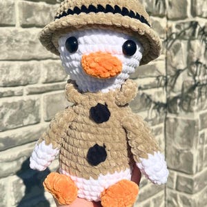 May include: A handmade crochet duck doll wearing a tan safari hat and jacket with black button accents. The duck has a white body, orange beak and feet, and black eyes. This plush toy is a unique, handcrafted item.