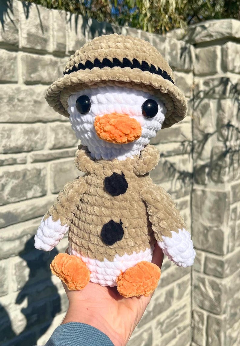 May include: A handmade crochet duck doll wearing a tan safari hat and jacket with black button accents. The duck has a white body, orange beak and feet, and black eyes. This plush toy is a unique, handcrafted item.