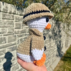 May include: A handmade crochet duck wearing a tan safari hat and jacket with navy blue accents. The duck has a white body, orange feet and beak, and black eyes. The plush toy is held in a person's hand.