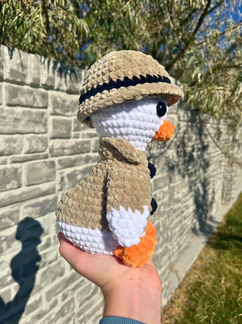May include: A handmade crochet duck wearing a tan safari hat and jacket with navy blue accents. The duck has a white body, orange feet and beak, and black eyes. The plush toy is held in a person's hand.