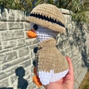 May include: A crocheted duck wearing a safari hat and jacket. The duck has a white body, orange beak and feet, and black eyes. The hat and jacket are tan with black and navy blue accents.