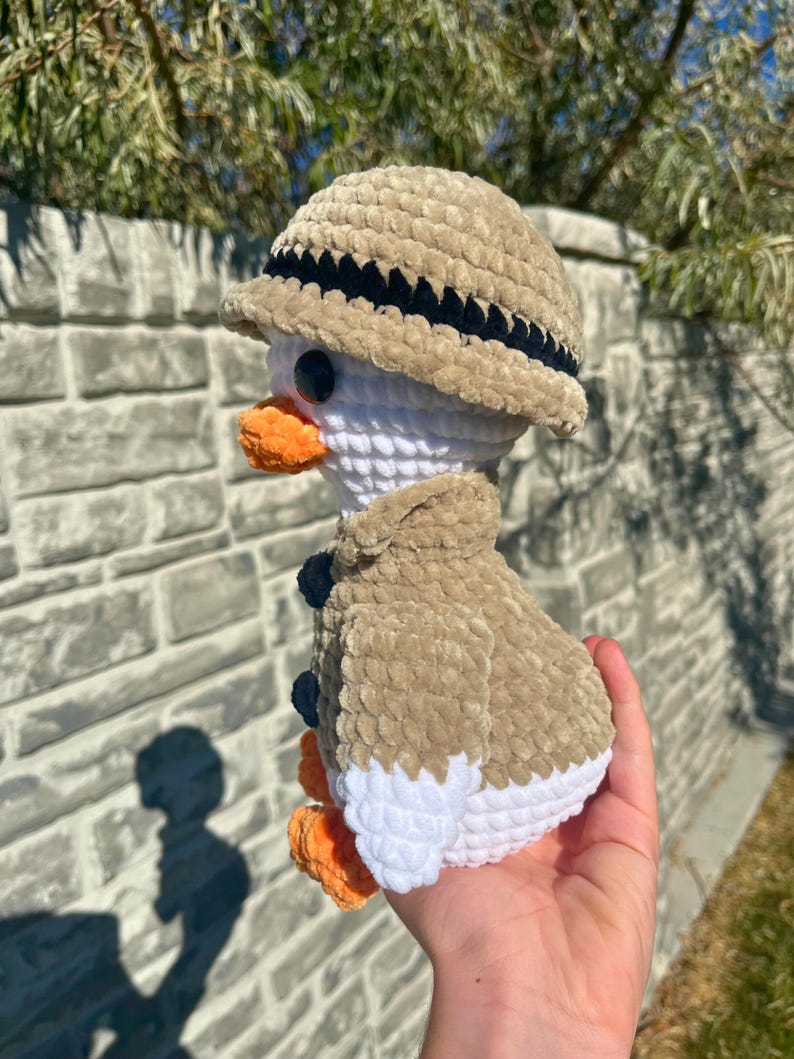 May include: A crocheted duck wearing a safari hat and jacket. The duck has a white body, orange beak and feet, and black eyes. The hat and jacket are tan with black and navy blue accents.