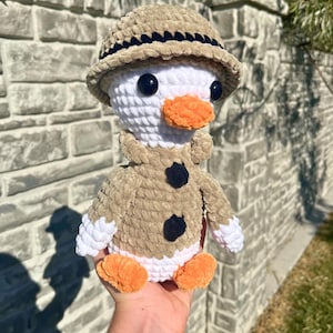 May include: A handmade crochet duck wearing a tan safari hat and jacket with black buttons. The duck has a white head, orange beak and feet, and black eyes. The plush toy is held in a hand.