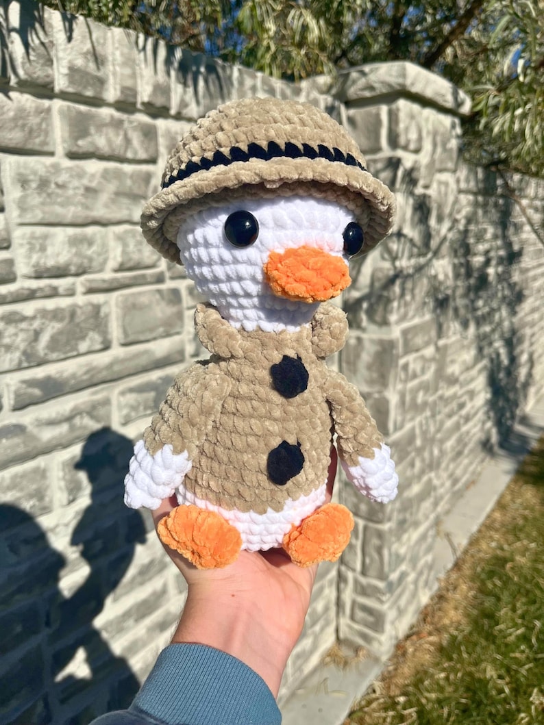 May include: A handmade crochet duck wearing a tan safari hat and jacket with black buttons. The duck has a white head, orange beak and feet, and black eyes. The plush toy is held in a hand.