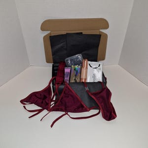 May include: A burgundy bikini set with a black box containing various items, including a small bottle of lotion, a pair of eyelash curlers, a small bottle of nail polish, and a pair of black tissue paper sheets.