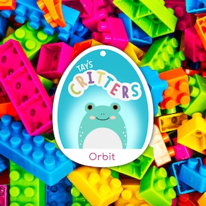May include: A blue and white egg-shaped tag with the text "Tay's Critters" and "Orbit" in a colorful font. The tag is surrounded by colorful plastic building blocks.