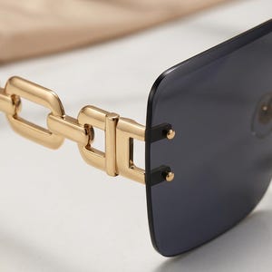 May include: Close-up of rimless sunglasses with a gold chain-link design on the temples. The lenses are a dark gray color, and the frame has a rectangular shape. The sunglasses are set against a neutral background.
