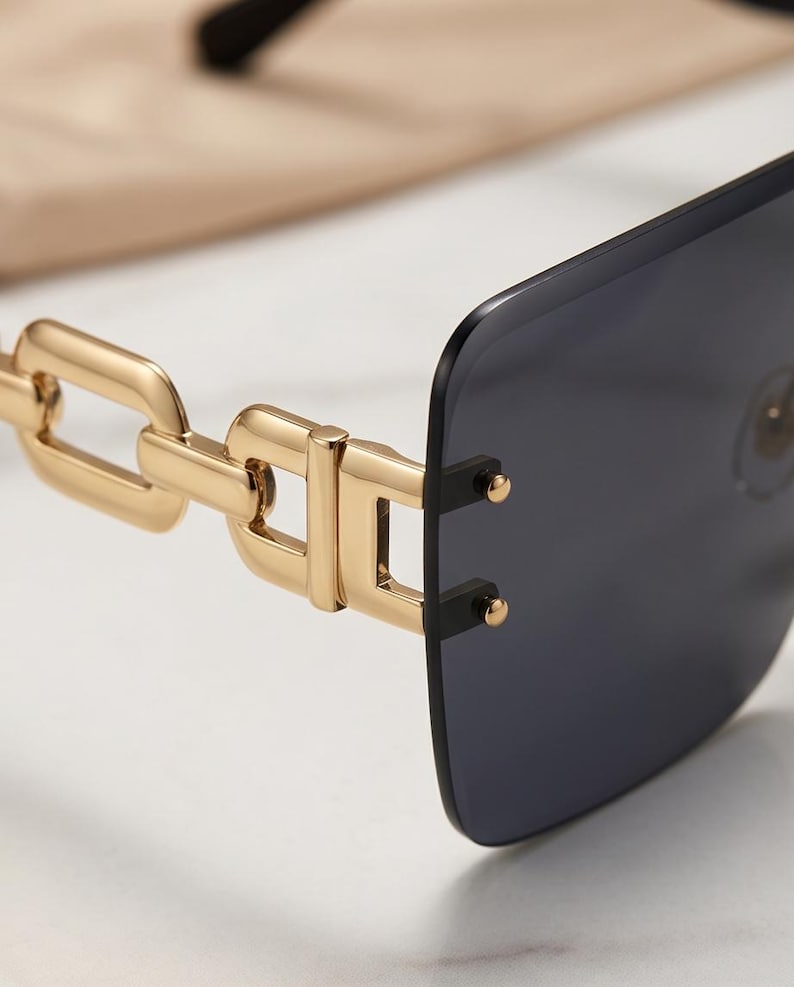 May include: Close-up of rimless sunglasses with a gold chain-link design on the temples. The lenses are a dark gray color, and the frame has a rectangular shape. The sunglasses are set against a neutral background.