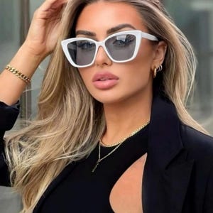 Pu&ograve; includere: Occhiali da sole bianchi a occhi di gatto con lenti scure. Gli occhiali da sole sono indossati con un top e un blazer neri. Sono visibili anche una collana e un braccialetto a catena d'oro.