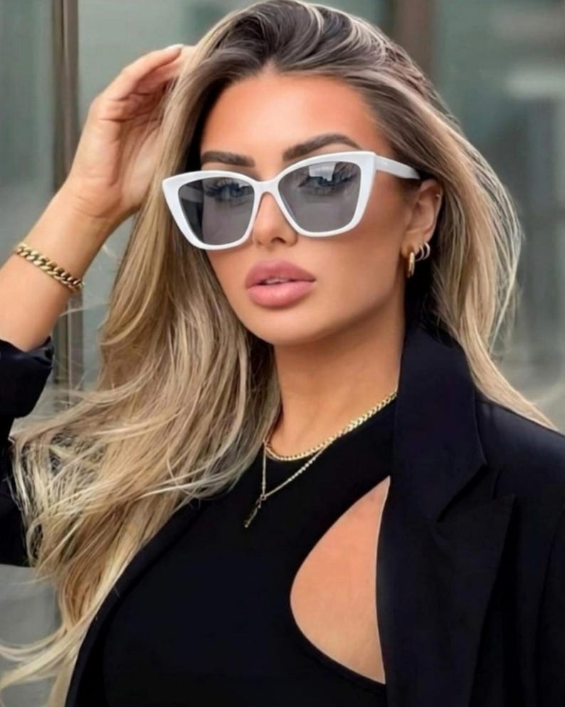 Pu&ograve; includere: Occhiali da sole bianchi a occhi di gatto con lenti scure. Gli occhiali da sole sono indossati con un top e un blazer neri. Sono visibili anche una collana e un braccialetto a catena d'oro.