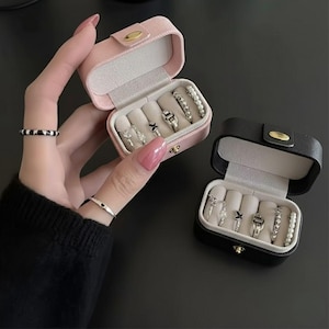 May include: A pink and black jewelry travel case with a gold clasp, open to reveal several rings. The rings are silver and feature various designs. The case is on a dark surface.