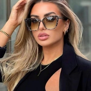 Pu&ograve; includere: Occhiali da sole quadrati con motivo leopardato sulle montature e lenti sfumate. Gli occhiali da sole sono indossati con un top e un blazer neri. La persona indossa anche gioielli d'oro, tra cui un braccialetto, una collana e orecchini.