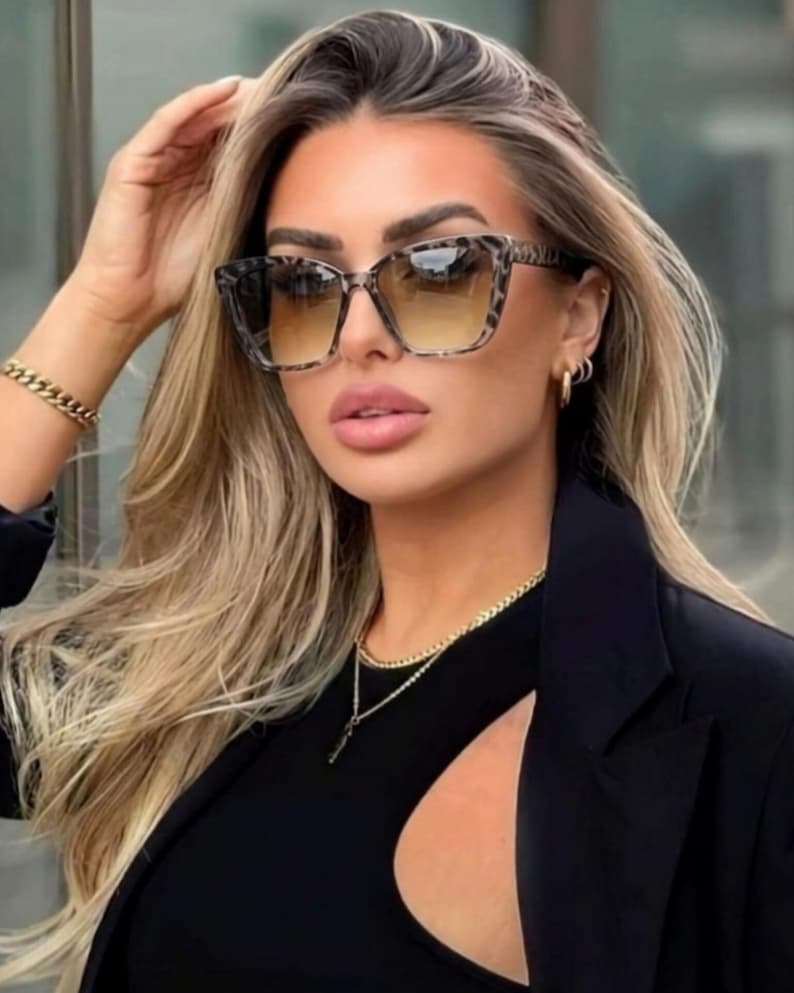 Pu&ograve; includere: Occhiali da sole quadrati con motivo leopardato sulle montature e lenti sfumate. Gli occhiali da sole sono indossati con un top e un blazer neri. La persona indossa anche gioielli d'oro, tra cui un braccialetto, una collana e orecchini.