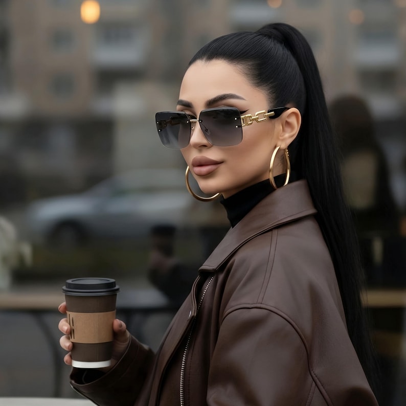 May include: Square-framed sunglasses with gradient lenses and gold accents. The sunglasses are paired with large gold hoop earrings. The person is wearing a brown leather jacket and holding a coffee cup.