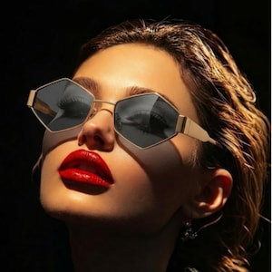 May include: Gold-framed sunglasses with a geometric, diamond-like shape. The lenses are dark, reflecting the surroundings. The image features a person with red lipstick, highlighting the sunglasses. The overall aesthetic is stylish and modern.