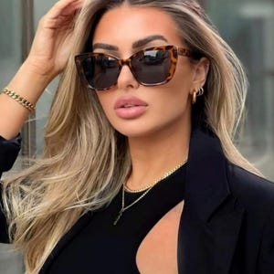 Pu&ograve; includere: Occhiali da sole con montatura tartarugata e lenti scure. Gli occhiali hanno una forma a occhi di gatto. La persona indossa un top nero e un blazer nero. Sono visibili anche una collana e un braccialetto a catena dorata.