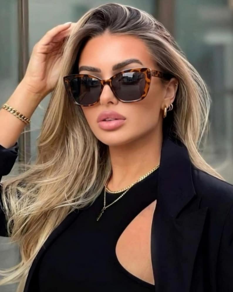 Pu&ograve; includere: Occhiali da sole con montatura tartarugata e lenti scure. Gli occhiali hanno una forma a occhi di gatto. La persona indossa un top nero e un blazer nero. Sono visibili anche una collana e un braccialetto a catena dorata.