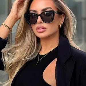 Pu&ograve; includere: Occhiali da sole neri a occhi di gatto con lenti scure. Gli occhiali sono indossati con un top nero, un blazer nero e gioielli dorati, tra cui una collana e un braccialetto. La persona ha i capelli biondi e indossa orecchini a cerchio dorati.