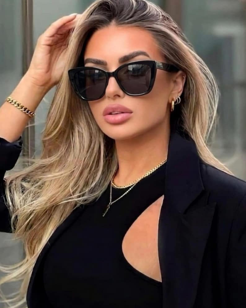 Pu&ograve; includere: Occhiali da sole neri a occhi di gatto con lenti scure. Gli occhiali sono indossati con un top nero, un blazer nero e gioielli dorati, tra cui una collana e un braccialetto. La persona ha i capelli biondi e indossa orecchini a cerchio dorati.