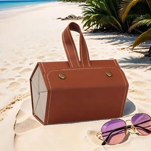 K&ouml;nnte beinhalten: Ein braunes Sonnenbrillenetui aus Kunstleder mit Griff und wei&szlig;en N&auml;hten, das an einem Sandstrand liegt. Goldgerahmte Sonnenbrillen mit violett-rosa Farbverlaufsgl&auml;sern liegen neben dem Etui. Das Etui hat zwei goldfarbene Druckkn&ouml;pfe.